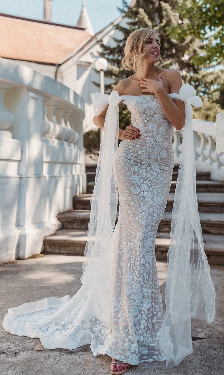 A woman wearing the Strapless White Wedding Gown: Tina Holly BB010, featuring sequins, floral-embroidered lace, and sheer ribbons, stands outdoors on stone steps near white railings and trees.